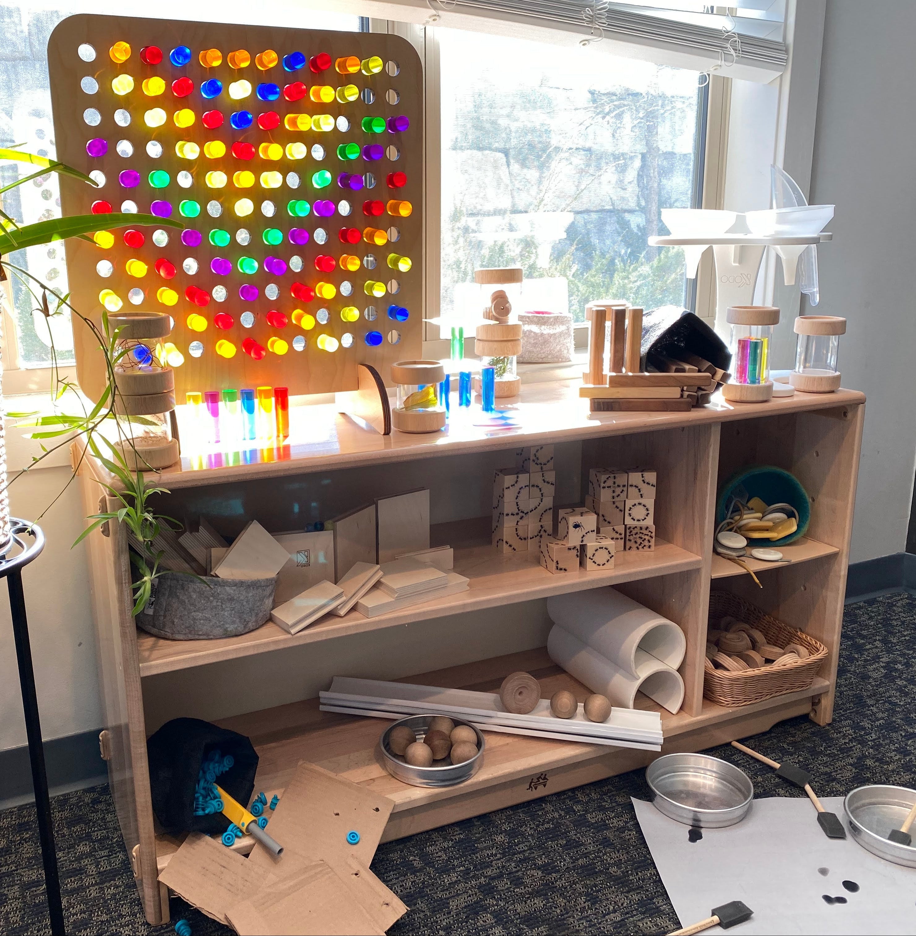 A wooden toy and art storage shelf in a bright indoor room, filled with colorful blocks, jars, rolls of paper, and a large illuminated multicolor circular light panel mounted on the back. A potted green plant sits on a black stand to the right, and a window is behind the shelf.
