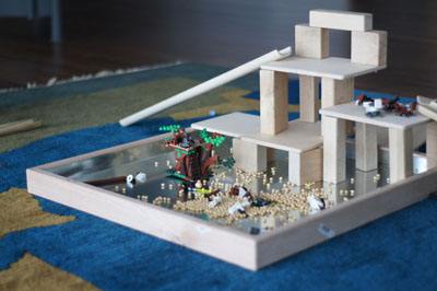 Maple wood activity tray with a reflective steel base, featuring a wooden block structure and small toys on a blue rug.