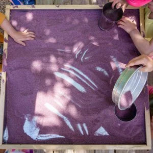Square maple activity tray with a reflective steel interior filled with purple sensory sand for children's play.