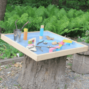 Maple wood activity tray with a reflective steel interior, set on a tree stump with colorful blocks and nature items.