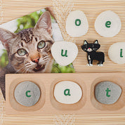 Engraved lowercase letter pebbles arranged on a wooden tray next to a cat photo and a small cat figurine.