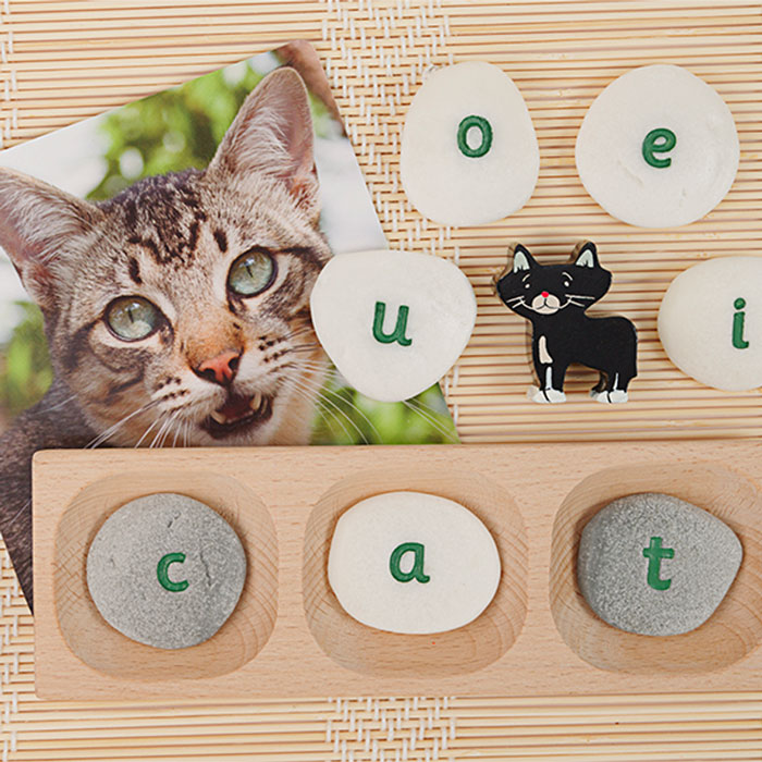 Engraved lowercase letter pebbles arranged on a wooden tray next to a cat photo and a small cat figurine.