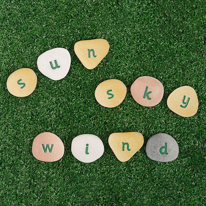Tactile, multi-colored stone and resin pebbles engraved with lowercase letters arranged on green artificial grass.