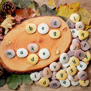 Tactile, multicolored stone and resin alphabet pebbles with engraved lowercase letters arranged on a wooden slice and autumn leaves.