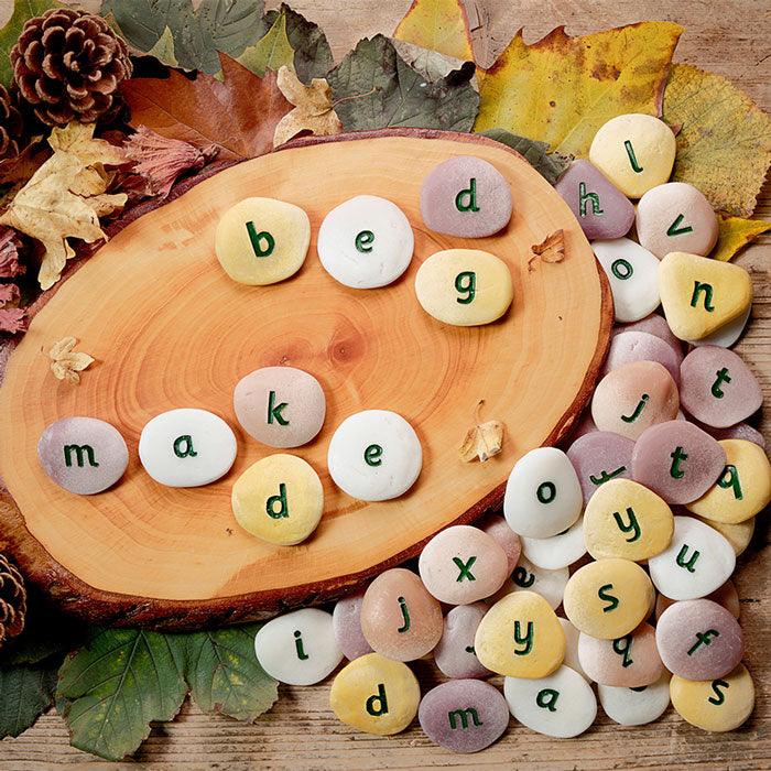 Tactile, multicolored stone and resin alphabet pebbles with engraved lowercase letters arranged on a wooden slice and autumn leaves.