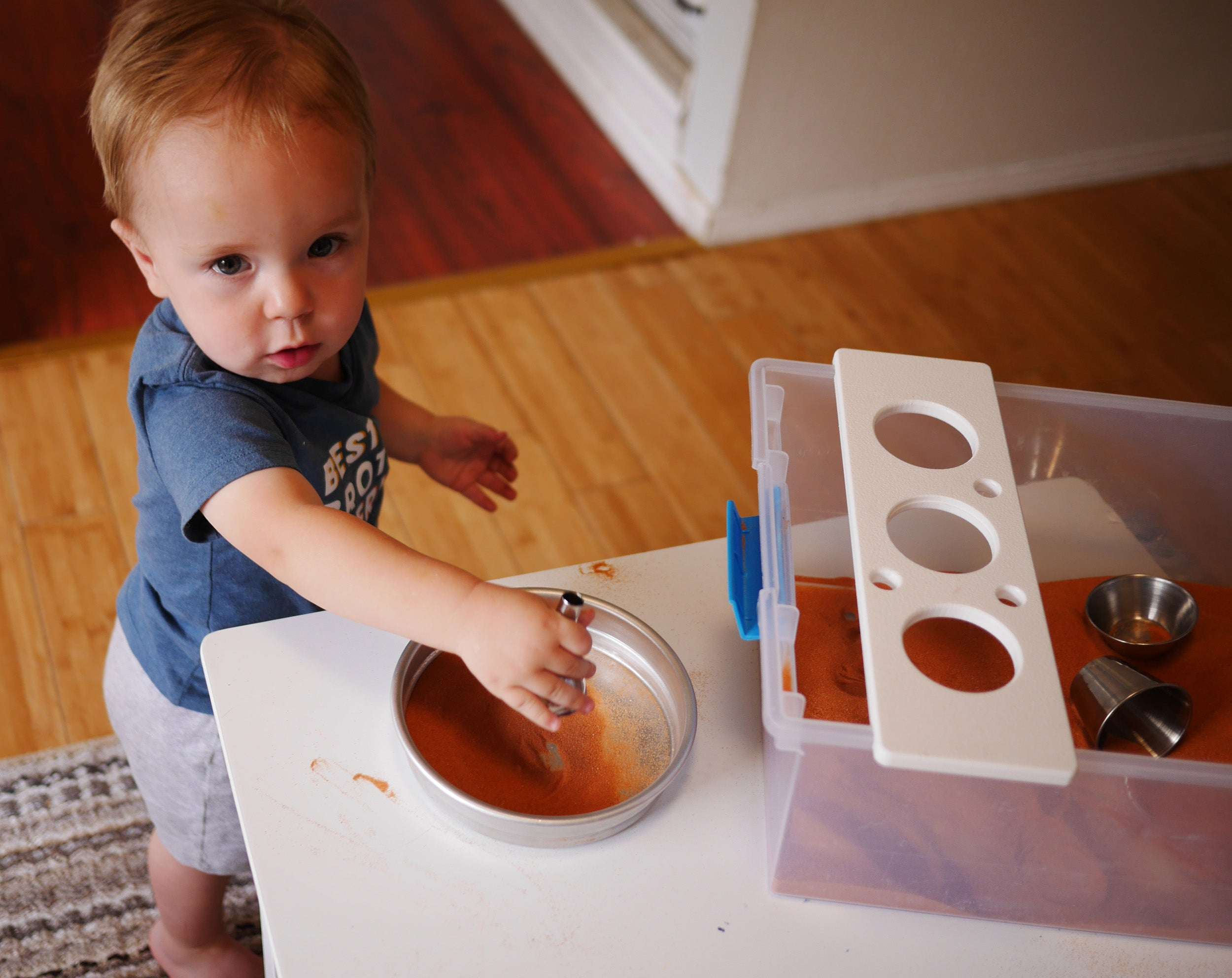 Toddler playing with a sensory bin containing orange Jurassic sand, metal cups, and a white tool holder on a white table.