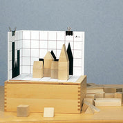 Wooden building blocks arranged on a grid board to match black shadow silhouettes on a white backdrop.