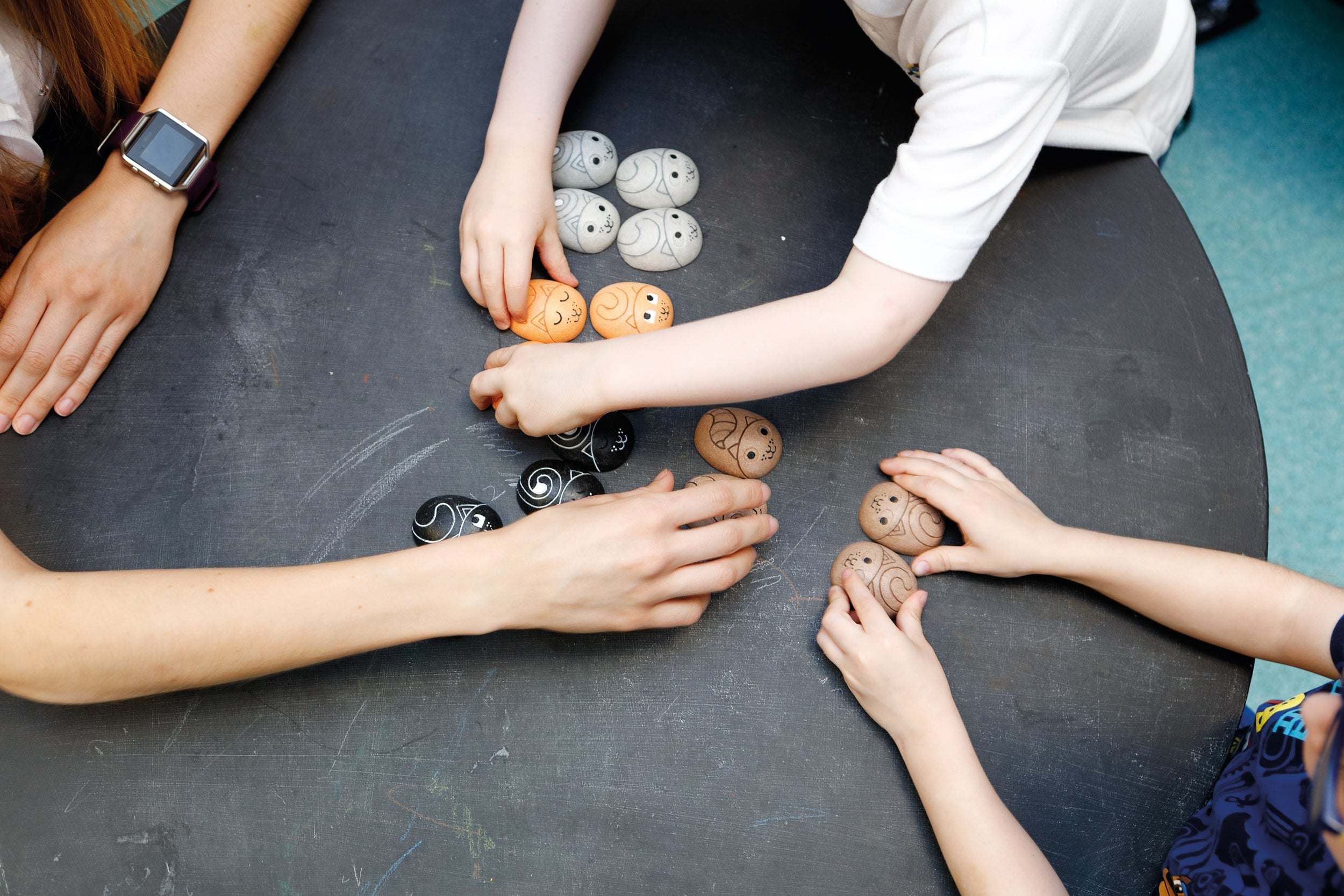 Hands sorting smooth, palm-sized CATegories attribute stones with engraved cat faces on a dark circular table.