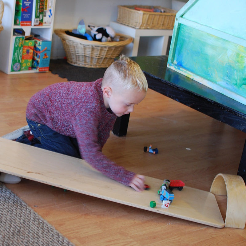 Child playing with a long, smooth wooden construction plank used as a ramp for toy cars on a classroom floor.