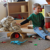 Child balancing a long wooden construction plank on an arch to create a seesaw with sandbags in a classroom.