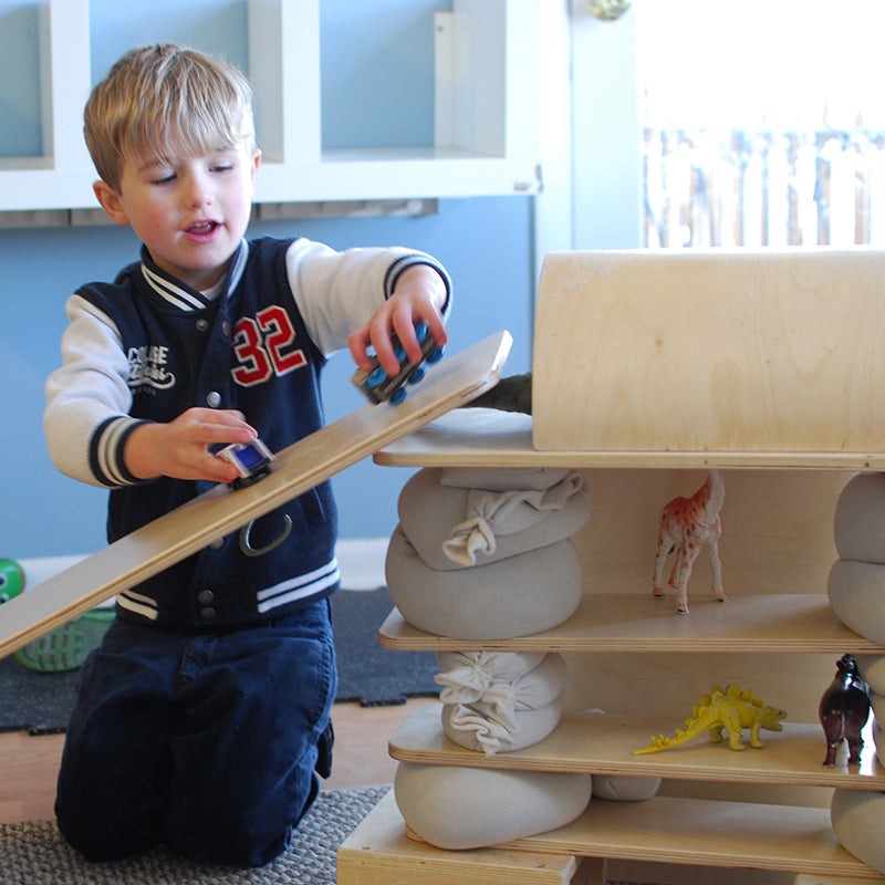 A young boy plays with a smooth, light-colored wooden construction plank as a ramp on a multi-level block structure.