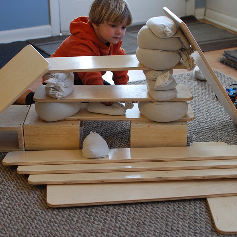 Child building with smooth, light-colored wooden construction planks and small canvas sandbags on a carpeted floor.