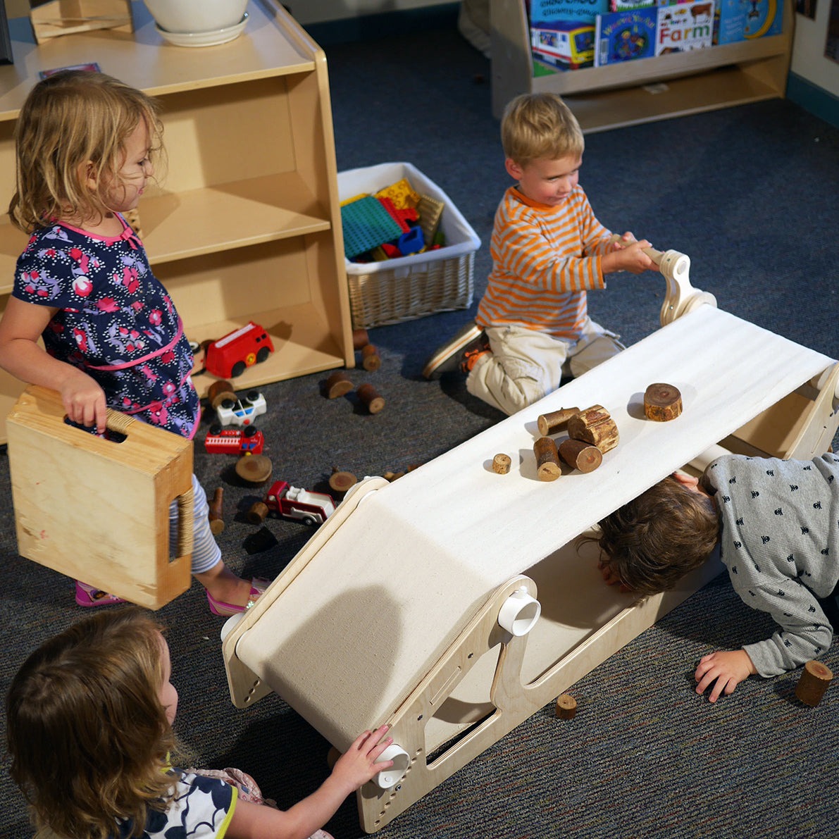 Children playing with a wooden conveyor belt toy featuring a white fabric belt, hand crank, and natural wood blocks.