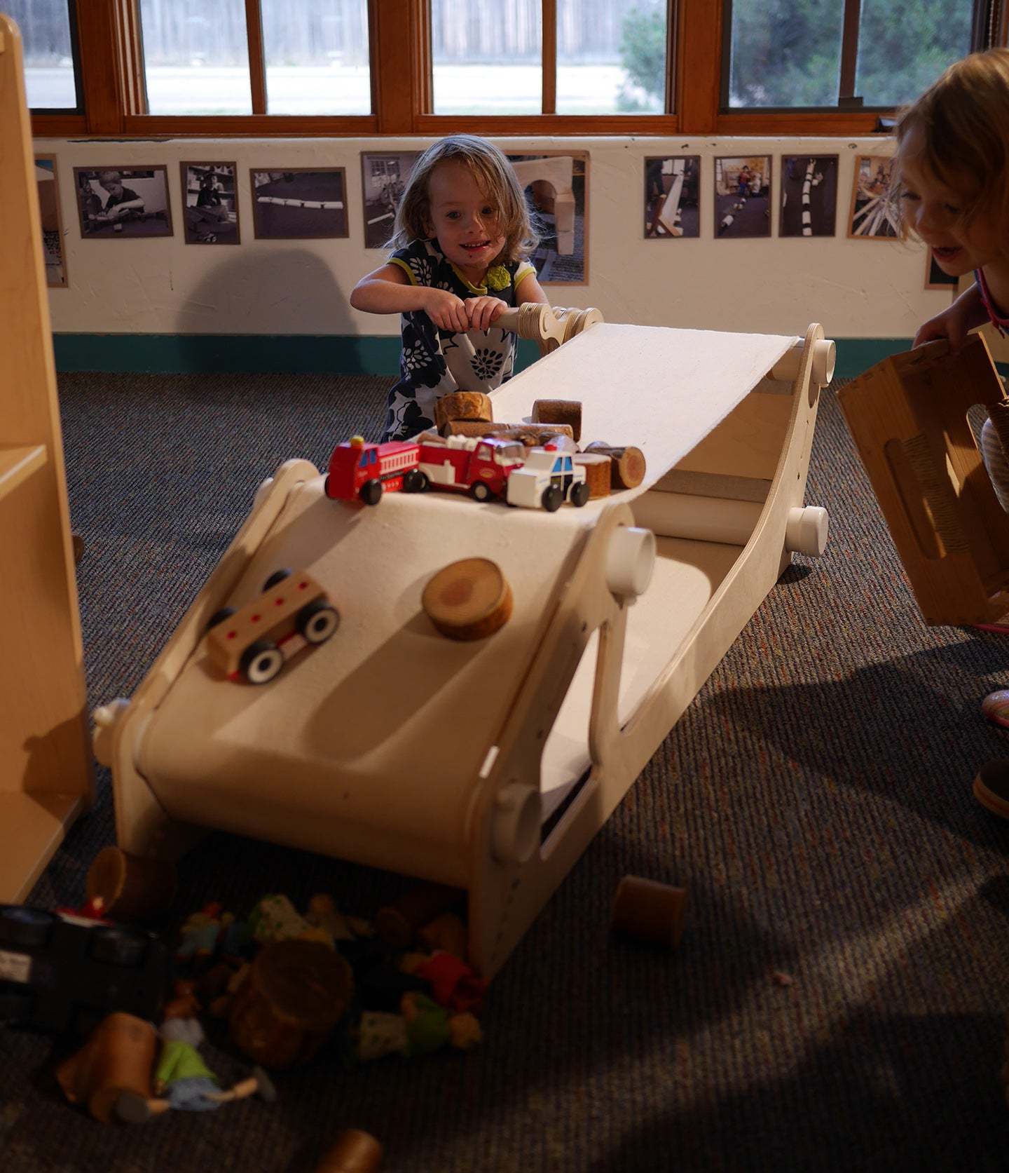 Child cranking a light wood conveyor belt toy with toy cars and blocks moving across the surface.