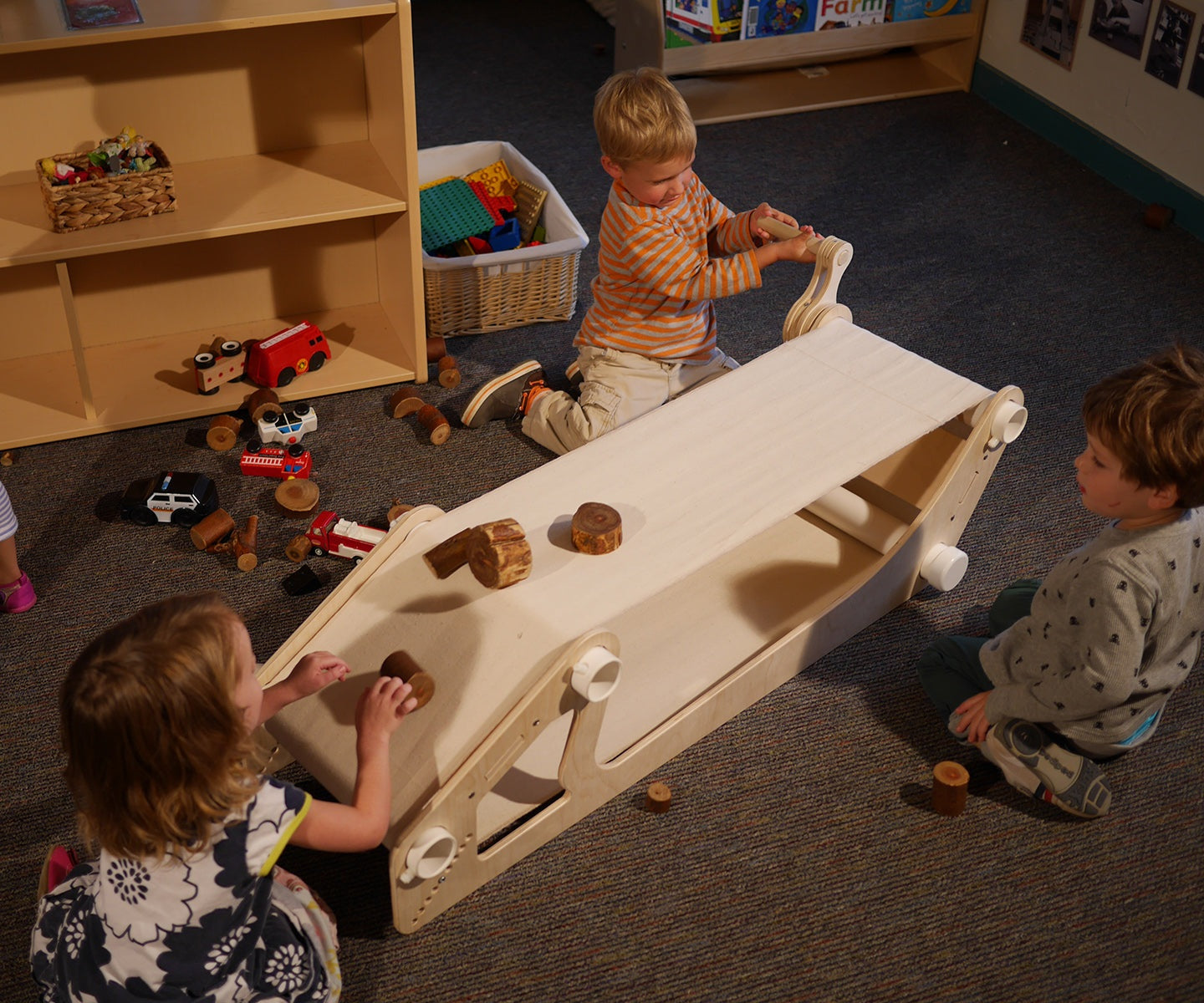Children playing with a wooden conveyor belt toy, cranking a handle to move small wooden blocks across the fabric surface.