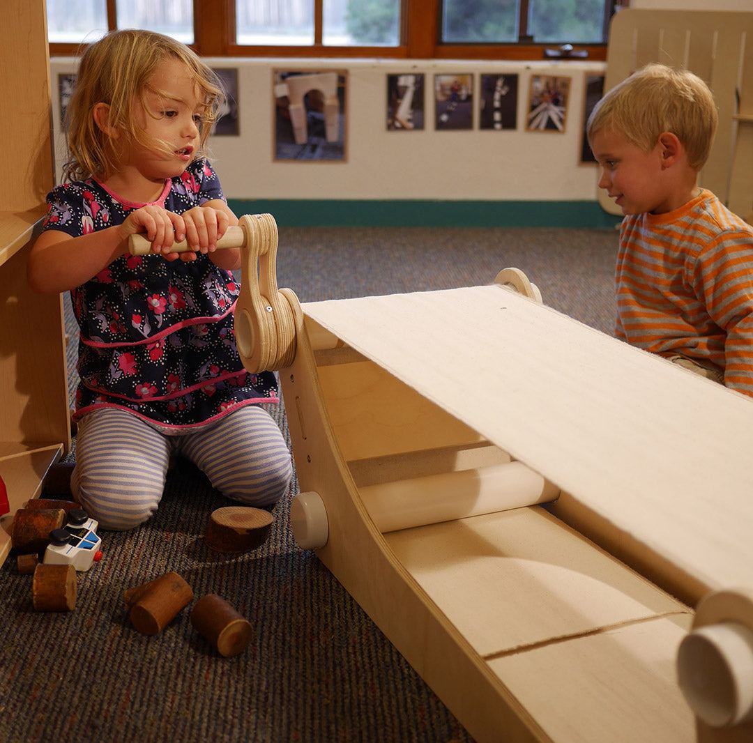 Child turning the handle of a light wood kinetic conveyor belt toy with a flat fabric surface and rollers.
