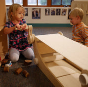 Child turning the handle of a light wood kinetic conveyor belt toy with a flat fabric surface and rollers.