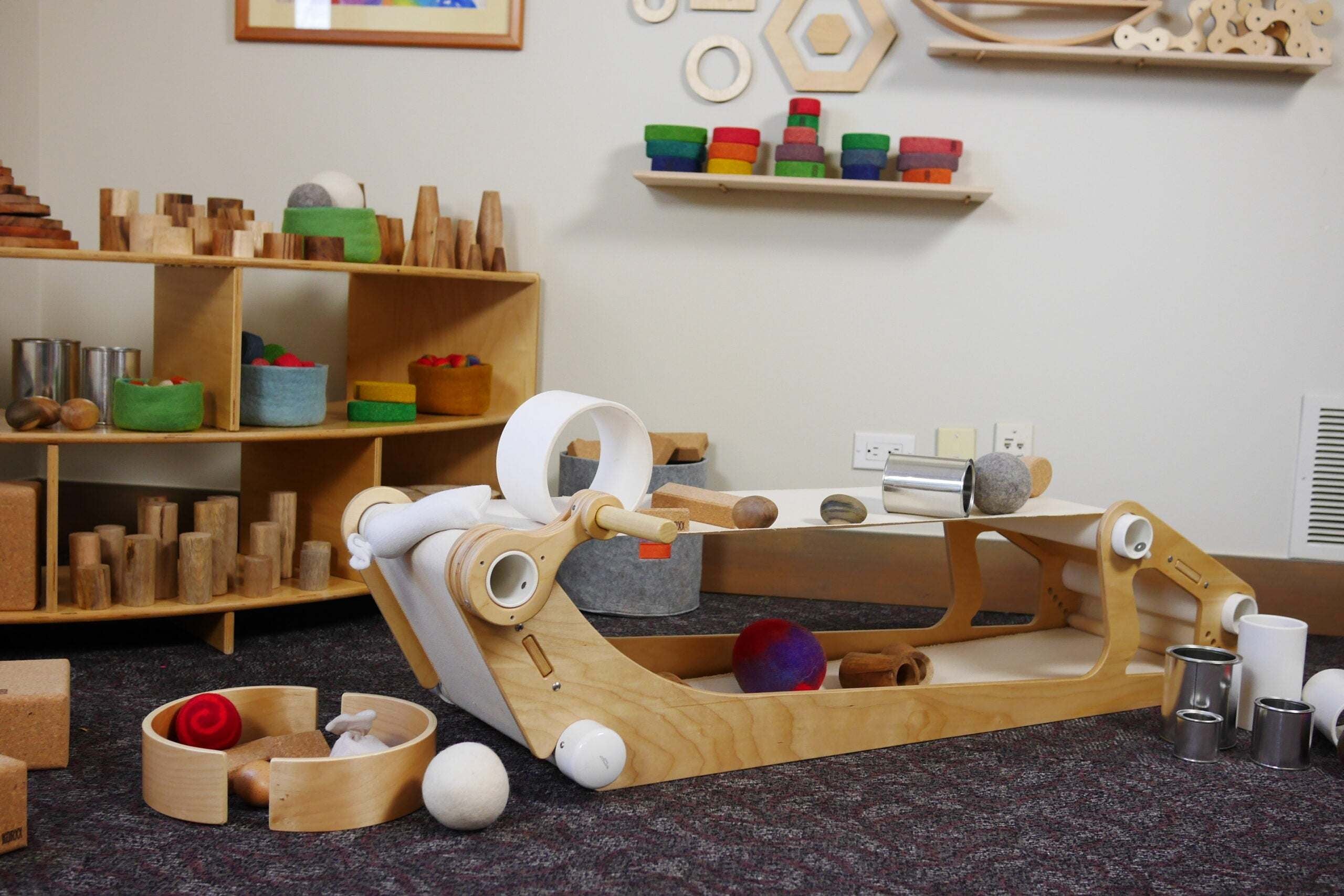Kid-powered wooden conveyor belt toy with a hand crank, white fabric belt, and various wooden and felt objects for kinetic play.