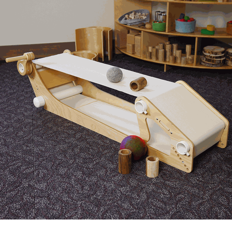 Kid-powered wooden conveyor belt toy with a hand crank, white fabric belt, and various wooden and felt play objects.
