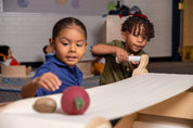 Two children playing with a wooden conveyor belt machine, turning a handle to move objects along a fabric track.