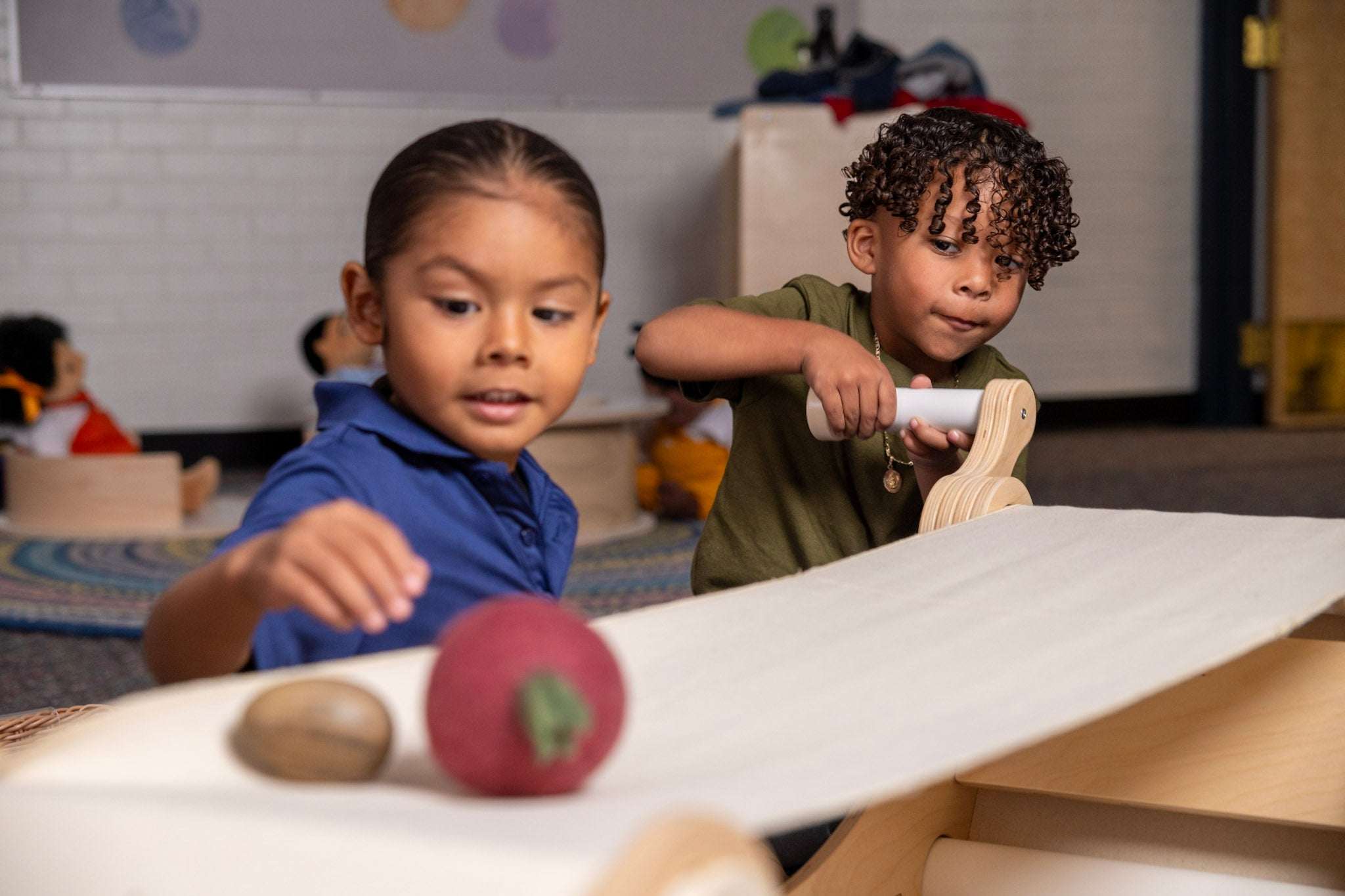 Two children playing with a wooden conveyor belt machine, turning a handle to move objects along a fabric track.