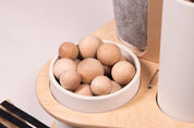 Natural wooden balls in a white ceramic bowl on a light birch wood caddy with a grey canvas storage pouch.