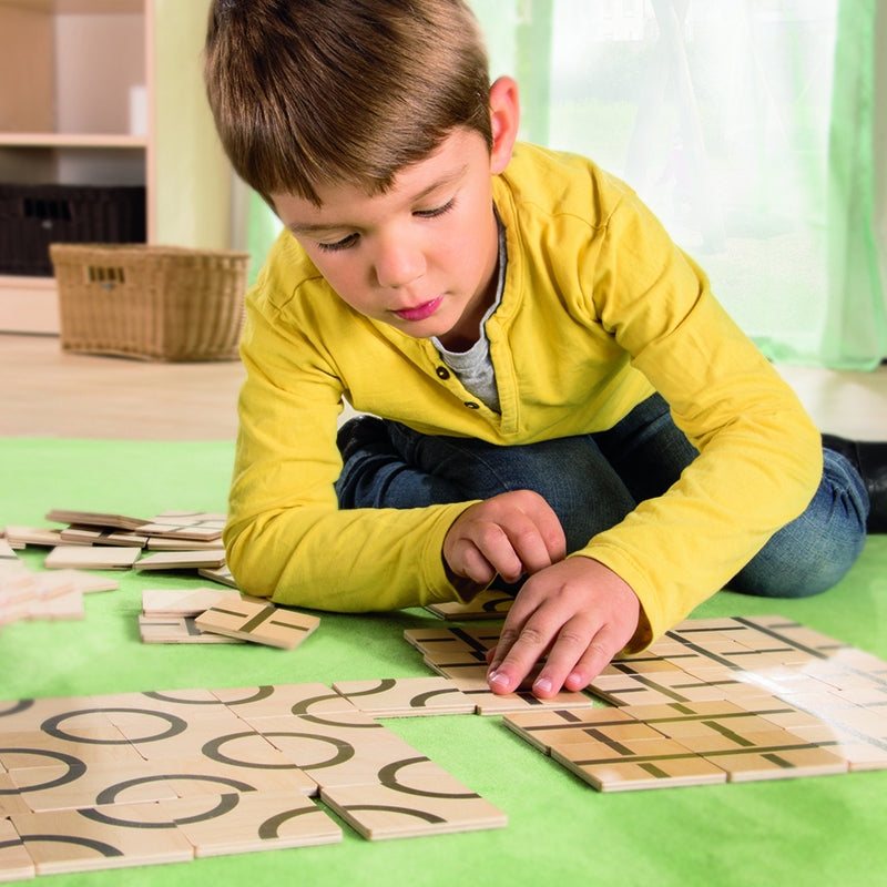 Child arranging square wooden tiles with black geometric patterns on a green mat.
