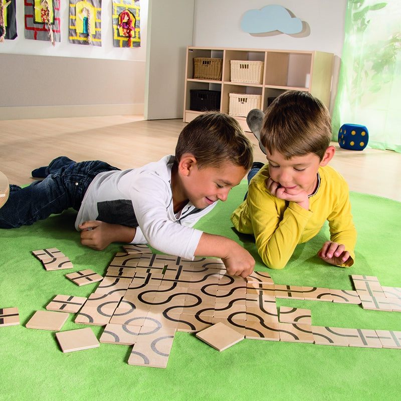 Two boys lying on a green rug arranging wooden square tiles with black printed curves and lines to create patterns.