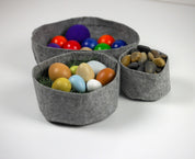 Three grey felt fabric bowls of varying sizes filled with colorful balls, smooth river stones, and decorative eggs.