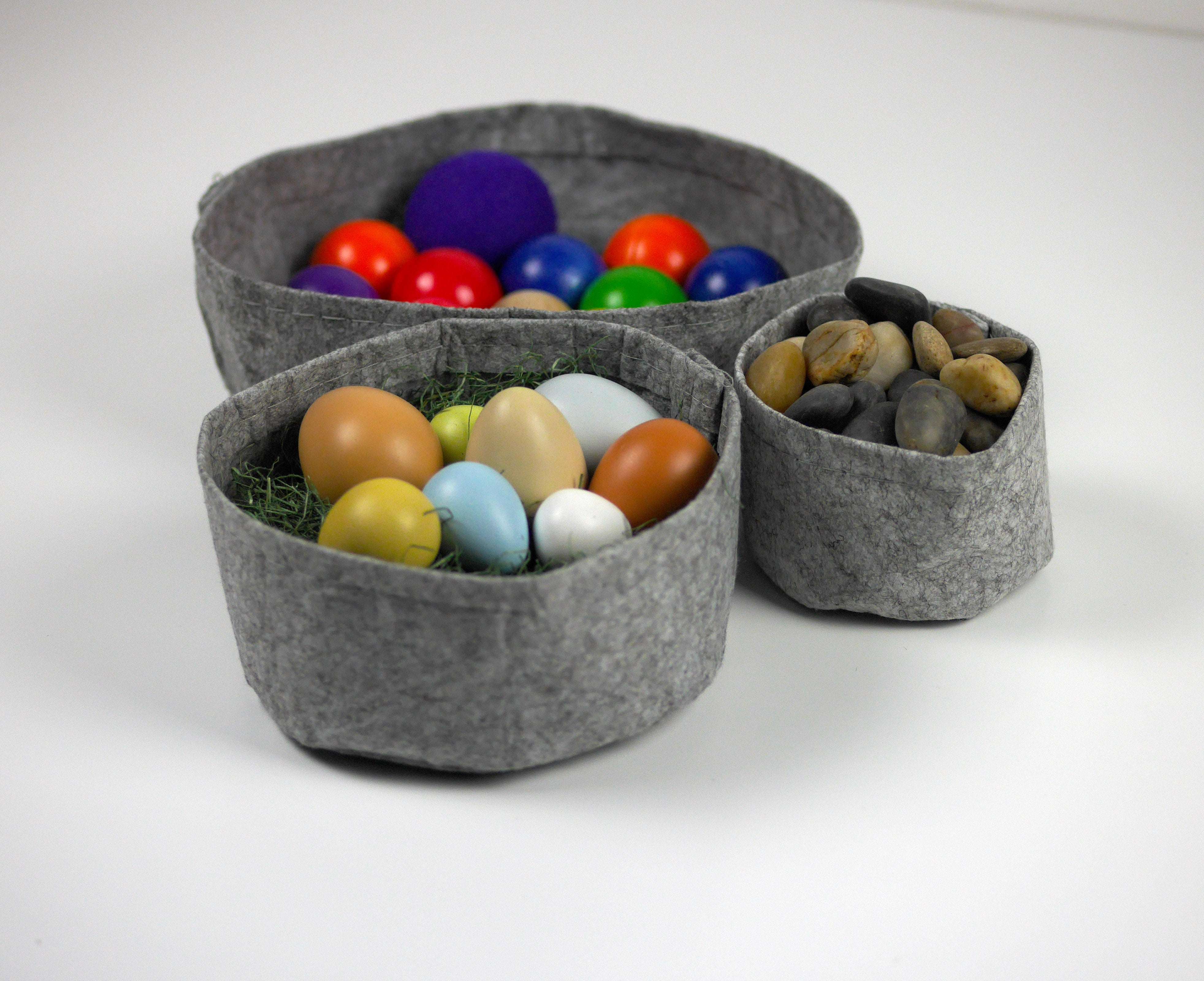 Three grey felt fabric bowls of varying sizes filled with colorful balls, smooth river stones, and decorative eggs.