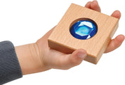 Child holding a chunky square wooden tile featuring a large, faceted blue gemstone insert.