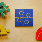 Four blue wooden jigsaw puzzle pieces assembled into a square on a light wood surface with colorful geometric shapes nearby.