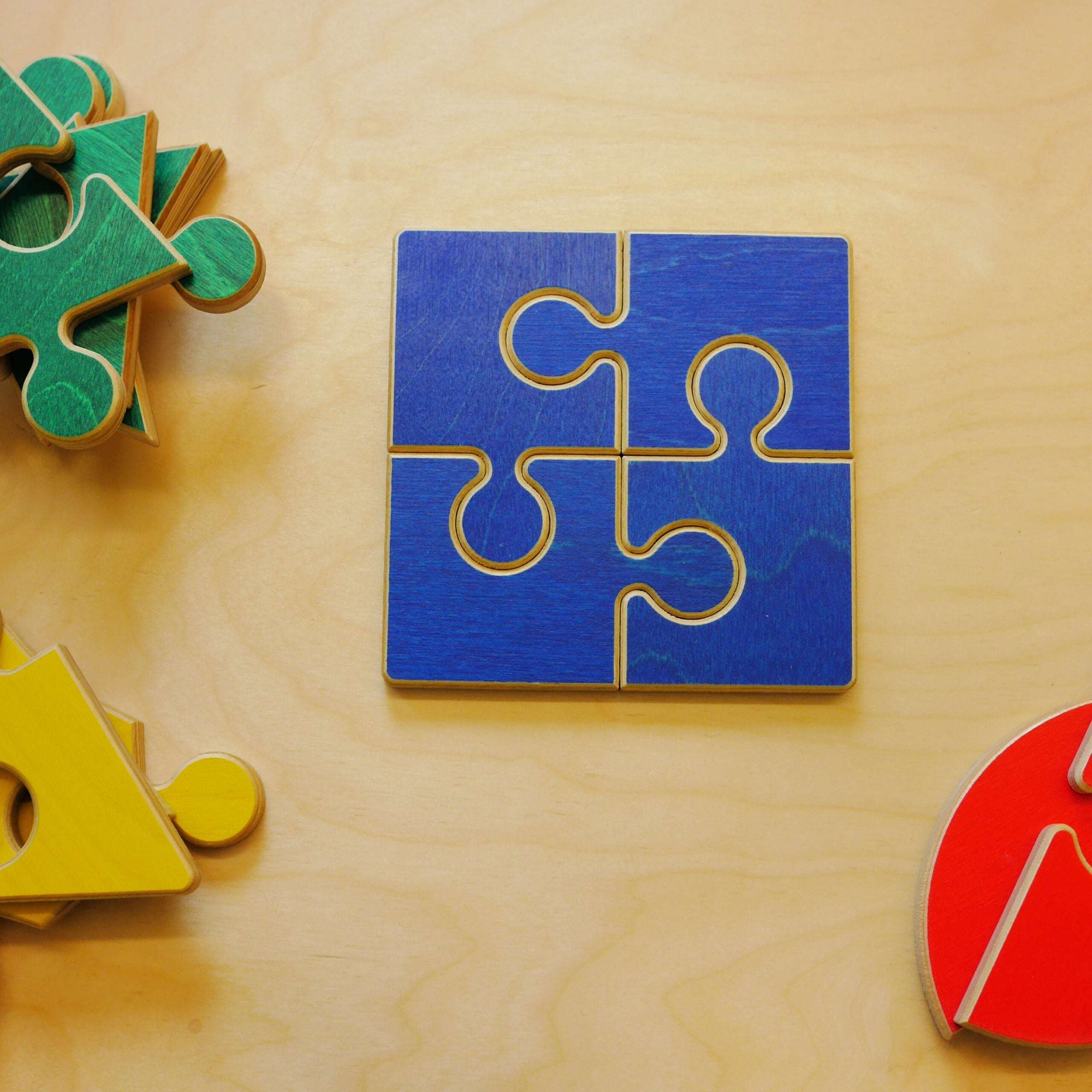 Four blue wooden jigsaw puzzle pieces assembled into a square on a light wood surface with colorful geometric shapes nearby.