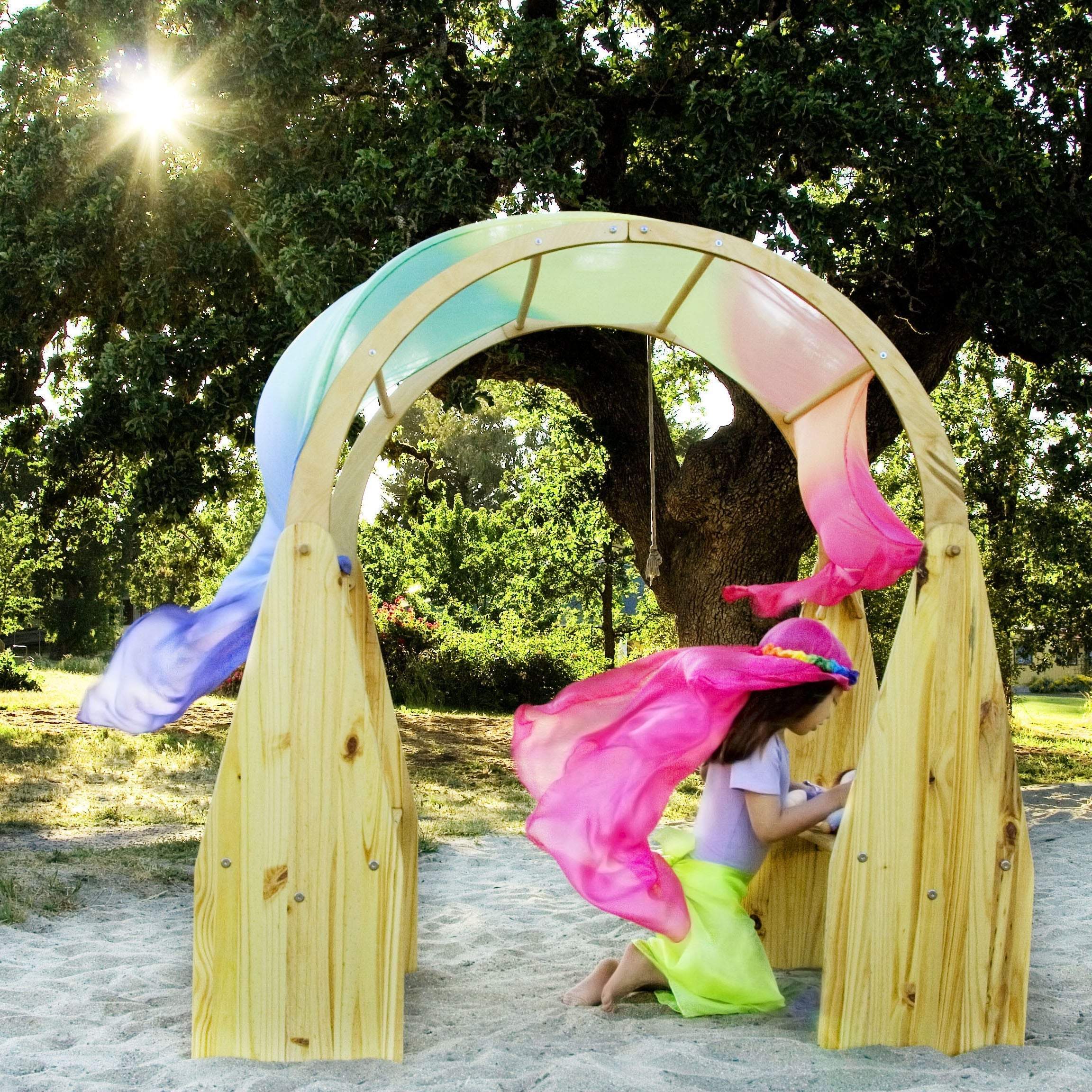 Child playing under a wooden arch draped with a giant, colorful rainbow silk scarf in a sunny outdoor setting.