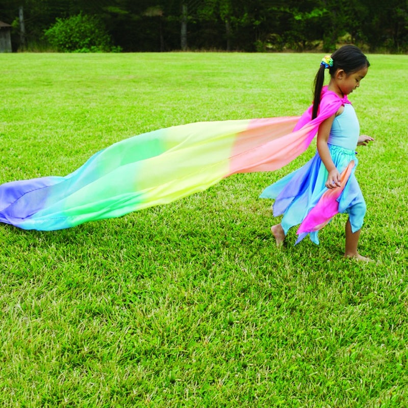 Child running in a grassy field wearing a flowing, giant rainbow-colored silk play cape.