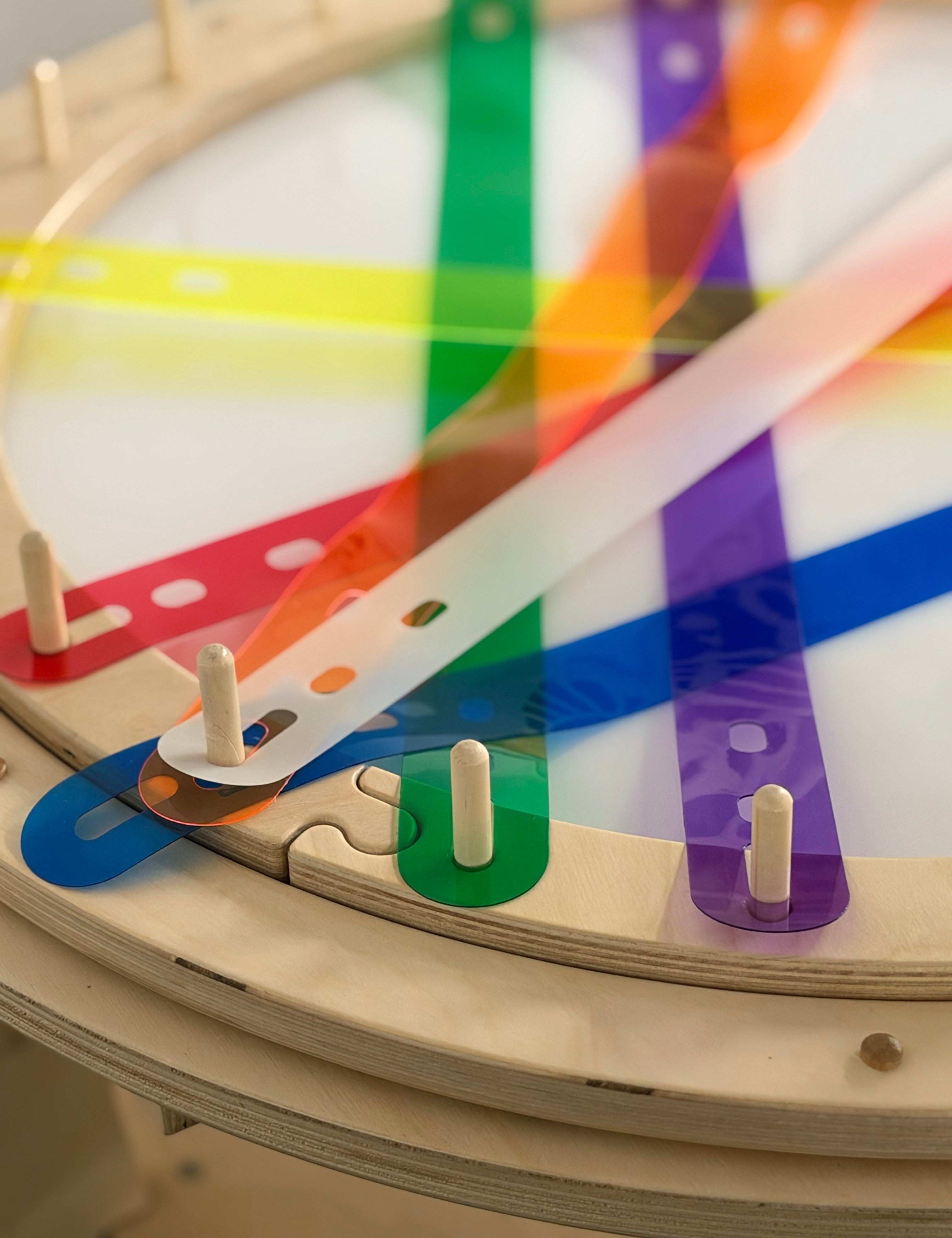 Round wooden weaving loom with colorful, translucent plastic strips attached to vertical pegs.