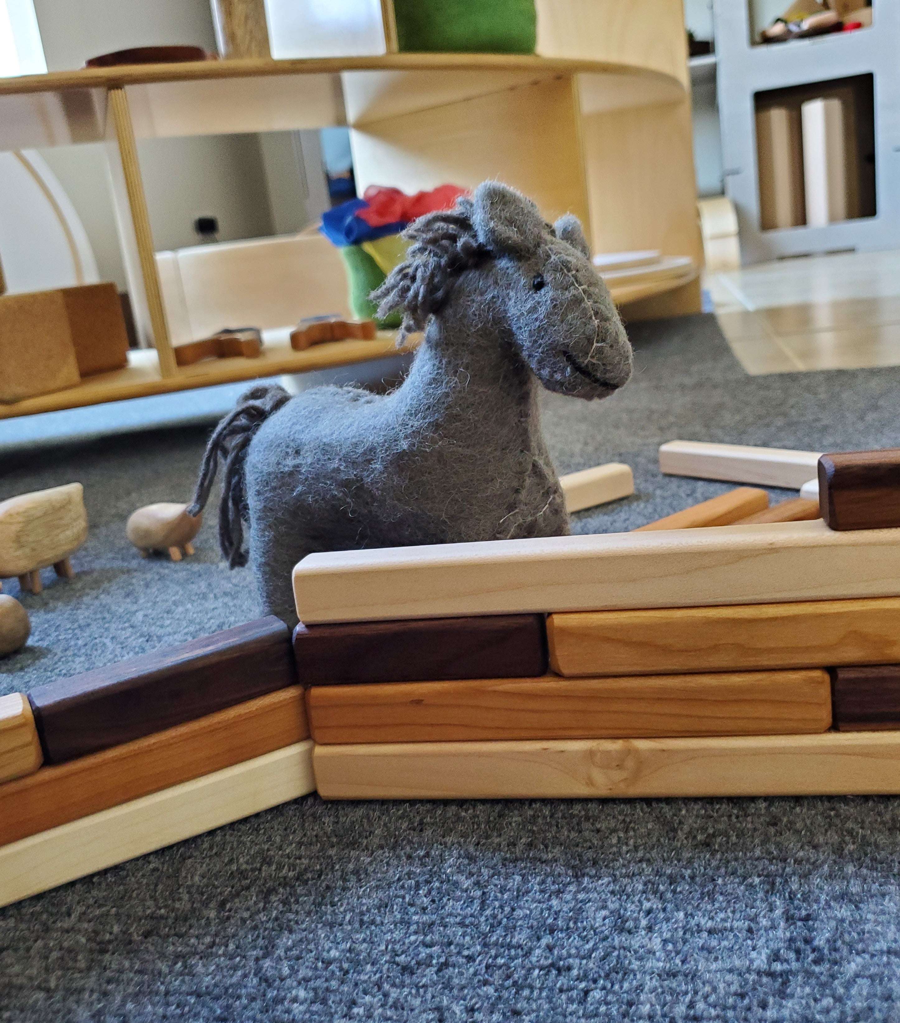 Felt grey horse toy standing behind a wall built from smooth, multi-toned wooden graduated building sticks.