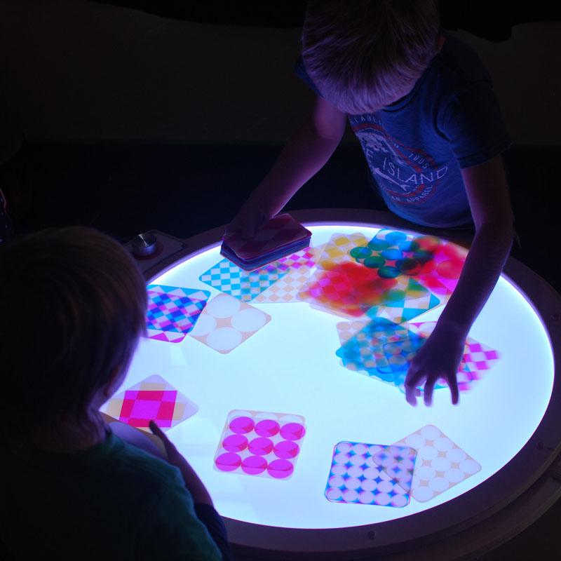 Children playing with colorful, transparent geometric cards on a glowing round light table to explore color mixing.