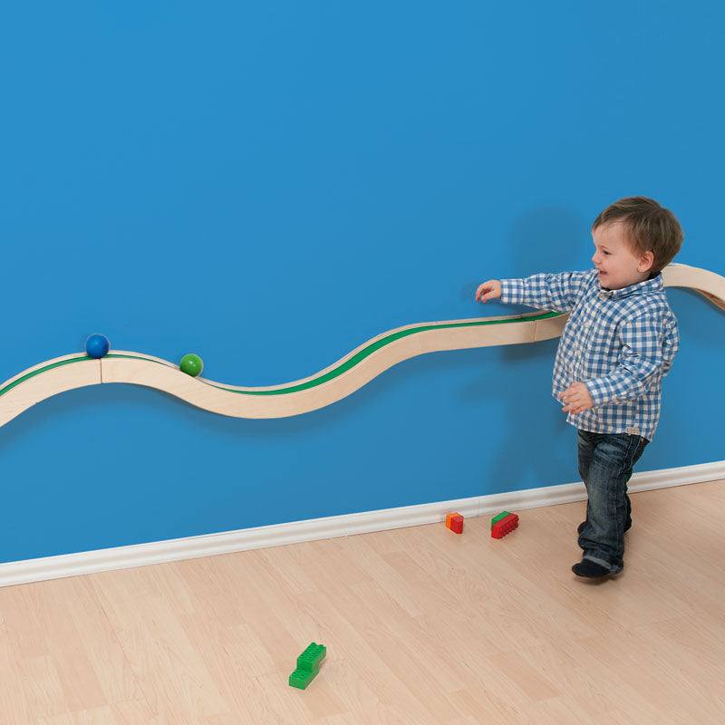 Toddler playing with a wooden Hill and Valley wall-mounted track featuring green paths and rolling balls on a blue wall.