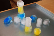 Set of four colorful, geometric ice molds used to create stacked blue, yellow, and clear ice shapes on a metal tray.