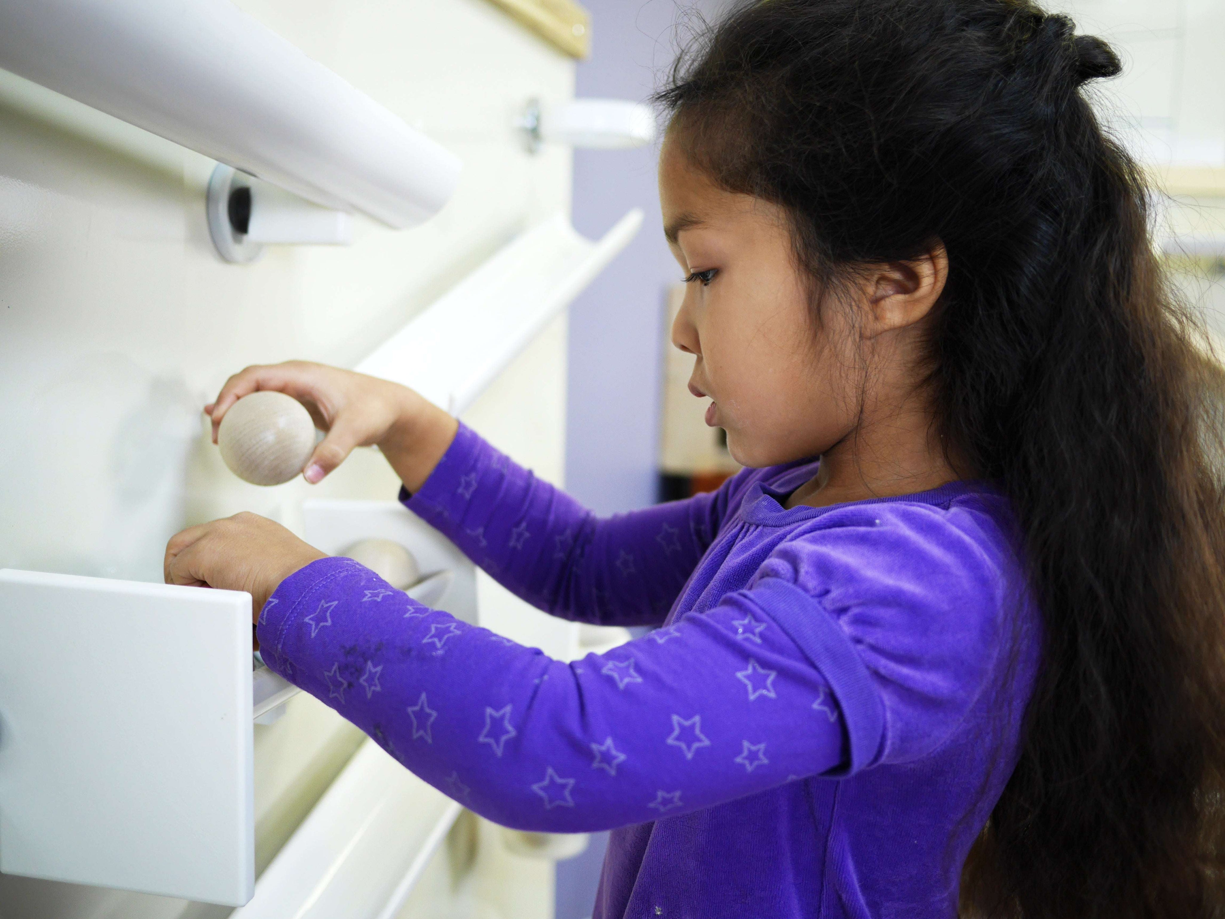 Girl in purple shirt playing with wooden balls on a white metal magnetic wall with ramps and Wormy Maple trim.