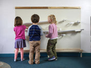 Three children playing with a large, wall-mounted magnetic STEM board featuring white metal panels and wormy maple wood trim.