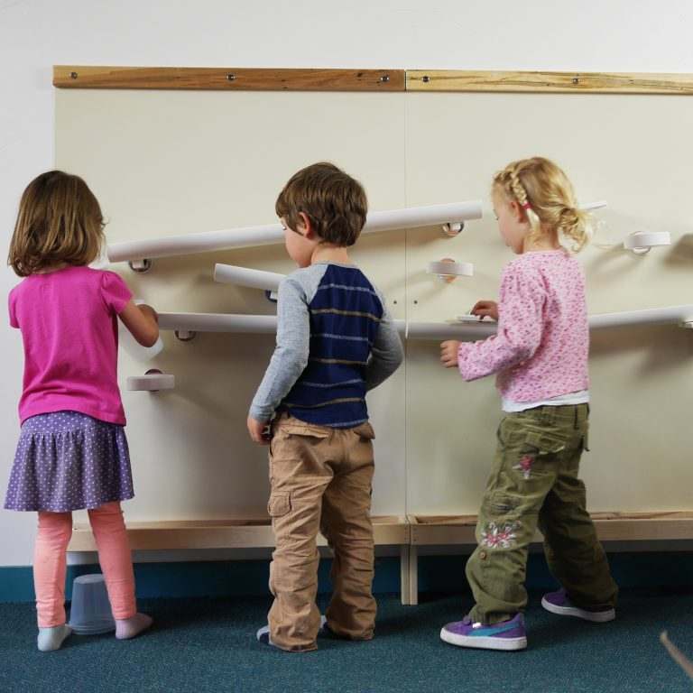 Children playing with magnetic ramps and cups on a large, cream-colored metal wall with durable Wormy Maple wood trim.