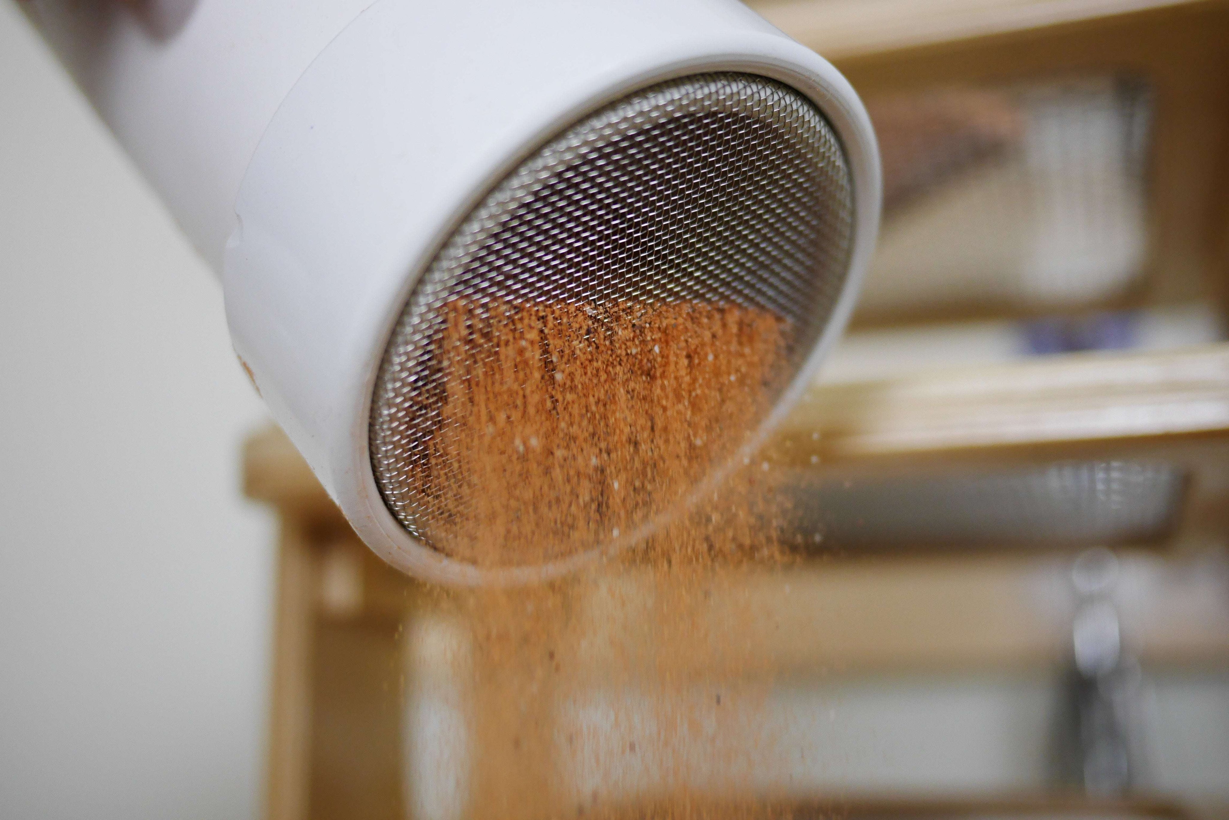 White plastic sifter scoop with a fine metal mesh screen pouring orange Jurassic sand.