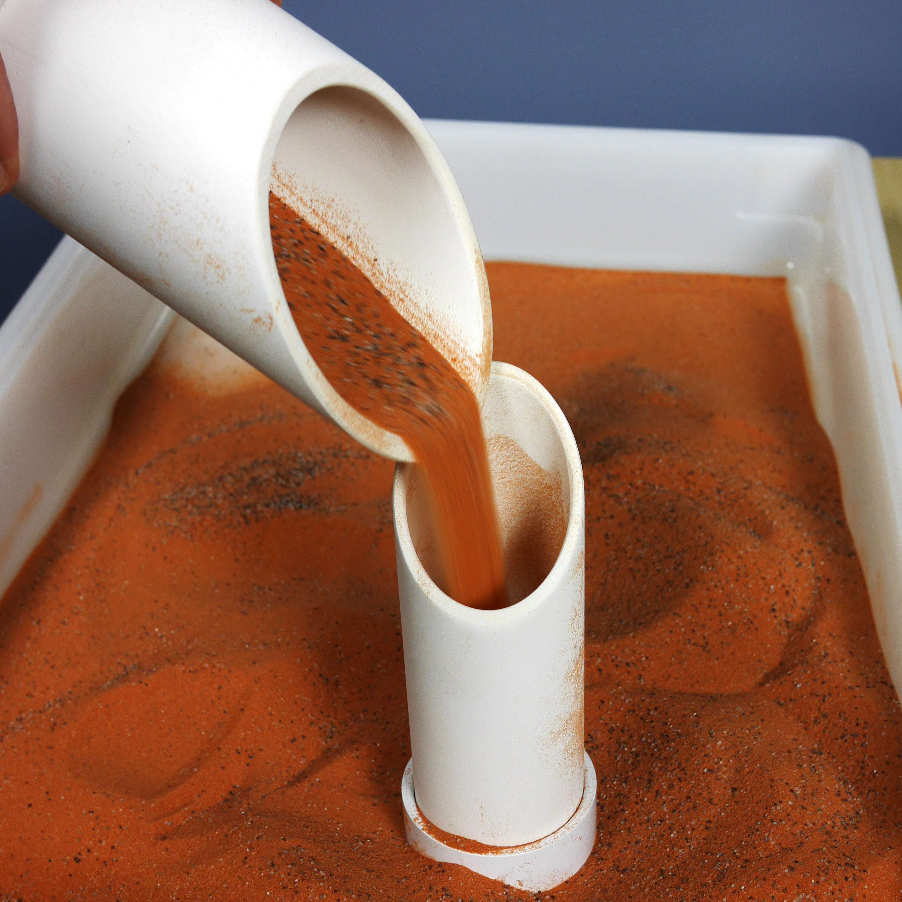 White plastic sifter scoops pouring orange Jurassic sand into a container for STEM play and treasure hunting.