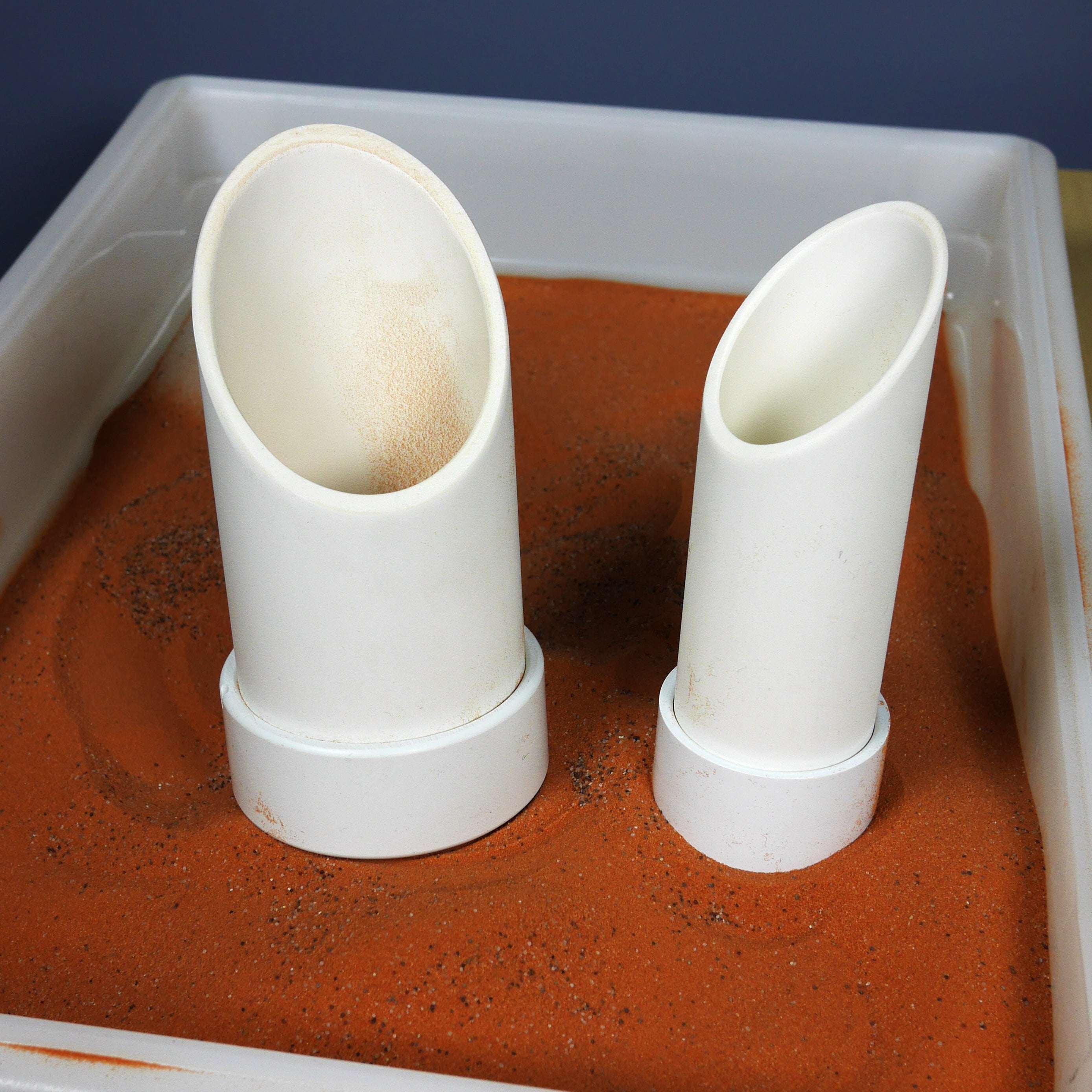 Two white, angled PVC sifter scoops standing in a white tray filled with fine, orange Jurassic sand.