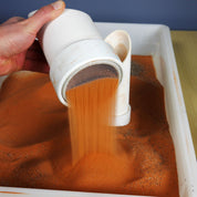 Hand holding a white plastic sifter scoop, straining fine orange sand into a white tray.