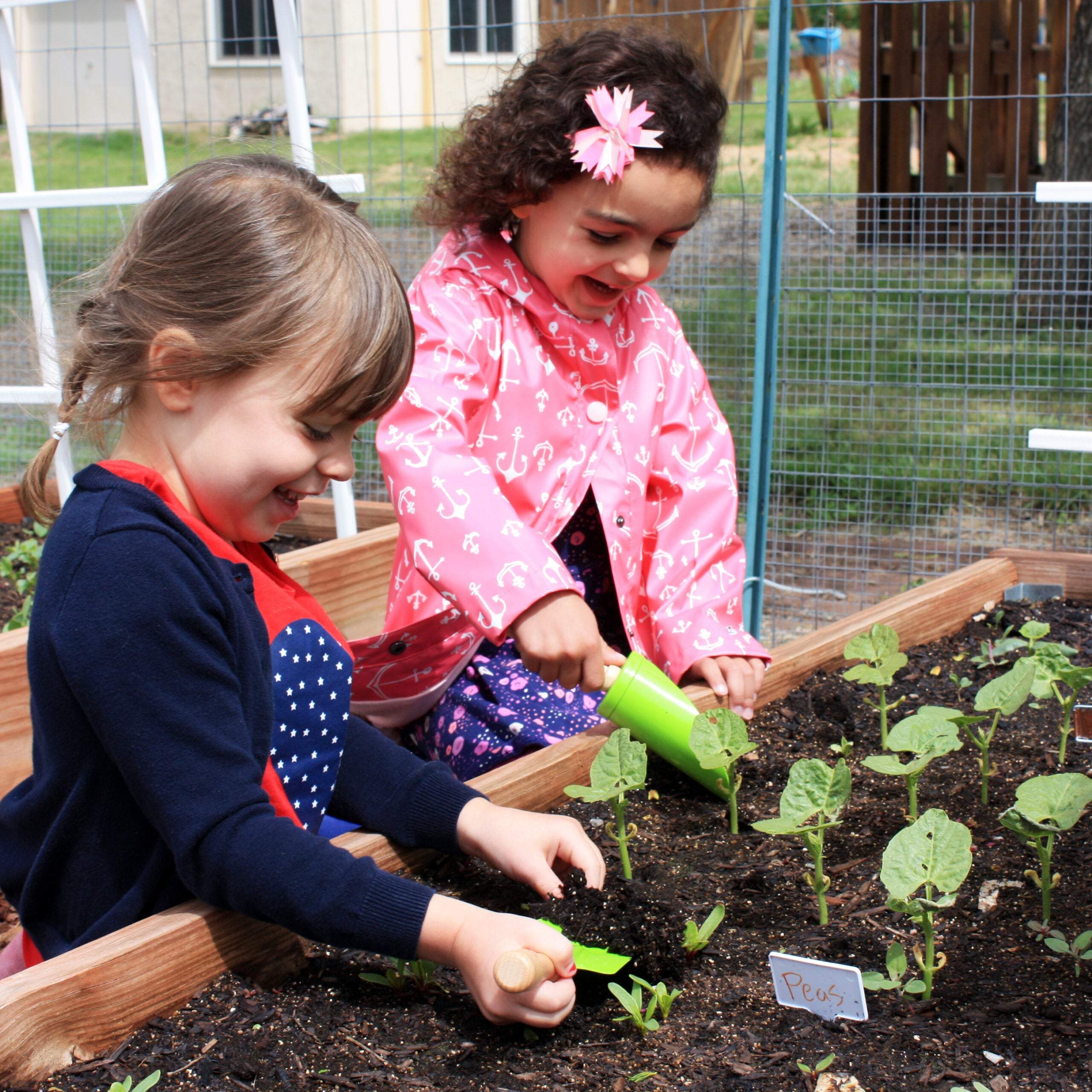 Two young girls using green garden hand tools to plant seedlings in a wooden raised garden bed.