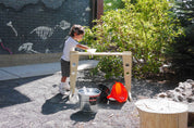 Child playing with a wooden funnel stand, metal bucket, and colorful plastic funnels in an outdoor setting.