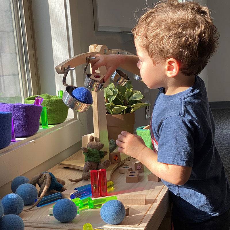 Child playing with a wooden balance scale, blue felt balls, and colorful gemstone tiles from the Kodo Basic Kit.